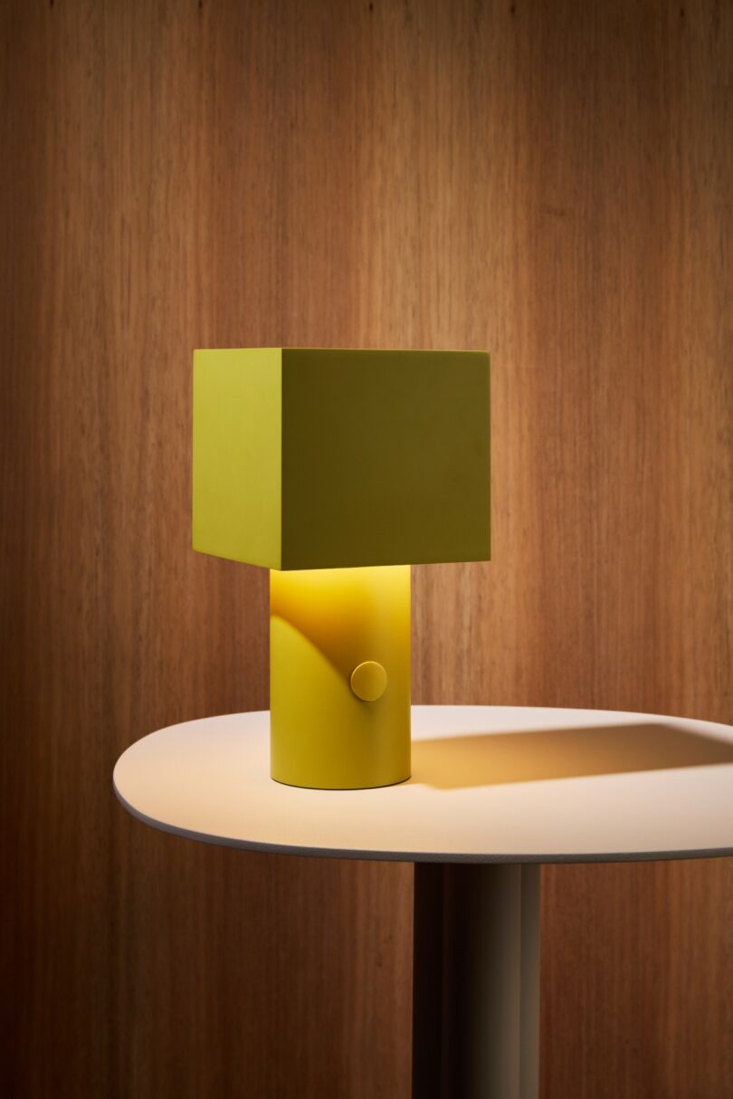 A yellow, modern table lamp with a square shade sits on a round, cream-colored table against a wooden background