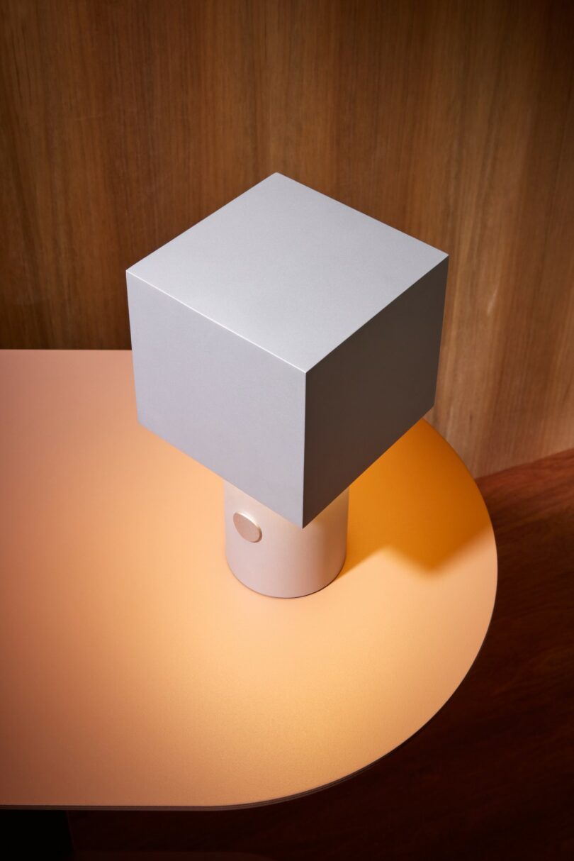 A white, cube-shaped lamp with a cylindrical base sits on a peach-colored table against a wooden background