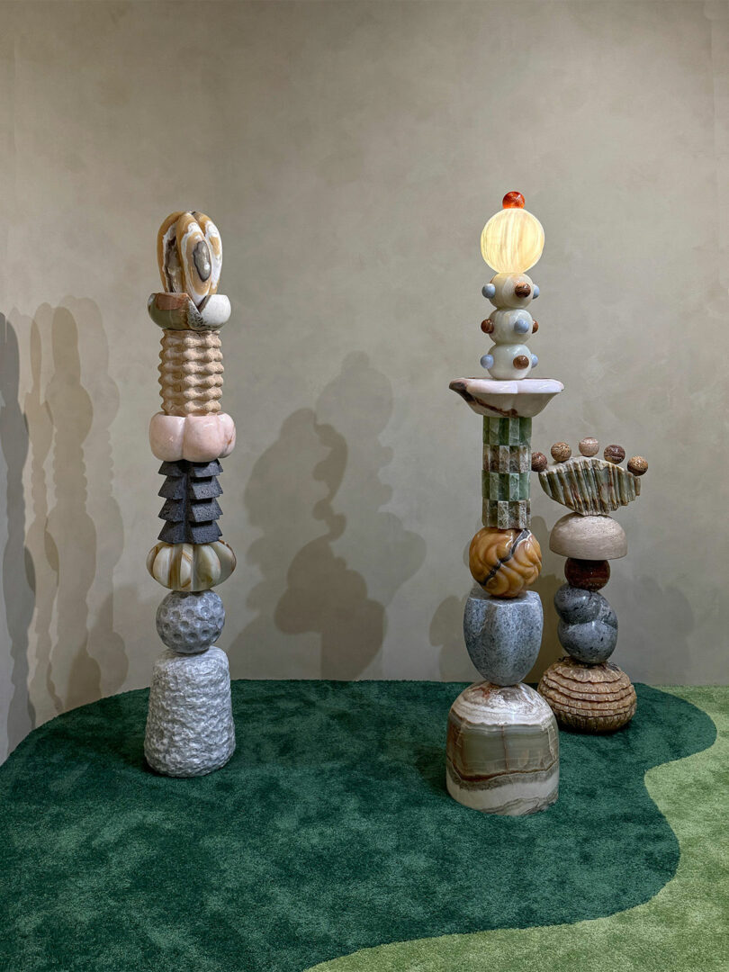 Three abstract, totem-like sculptures made of stacked, variously textured and colored shapes stand on a green and beige carpeted floor against a neutral wall.