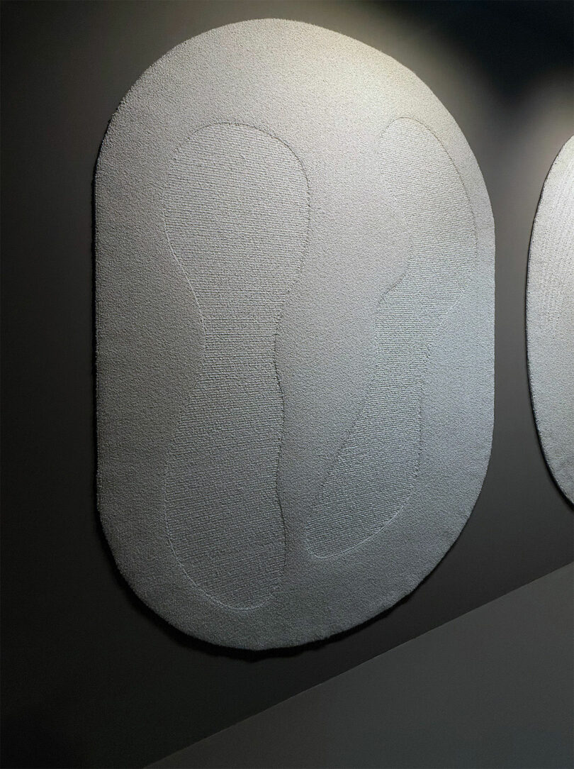 A textured, abstract wall hanging in an off-white color with faint curving lines, mounted on a dark grey wall under overhead lighting.