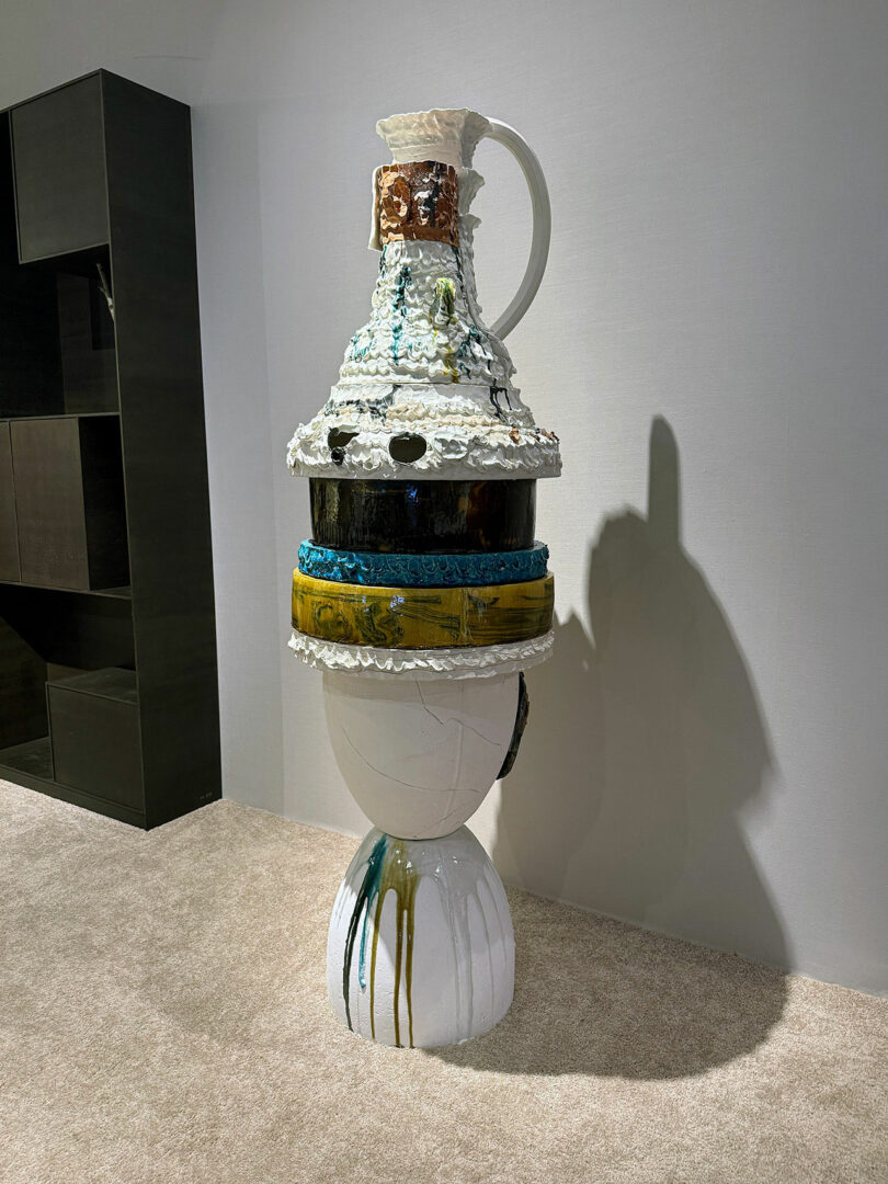 A tall, abstract ceramic sculpture with layered, colorful sections stands on a beige carpet against a plain wall, casting a shadow to the right.
