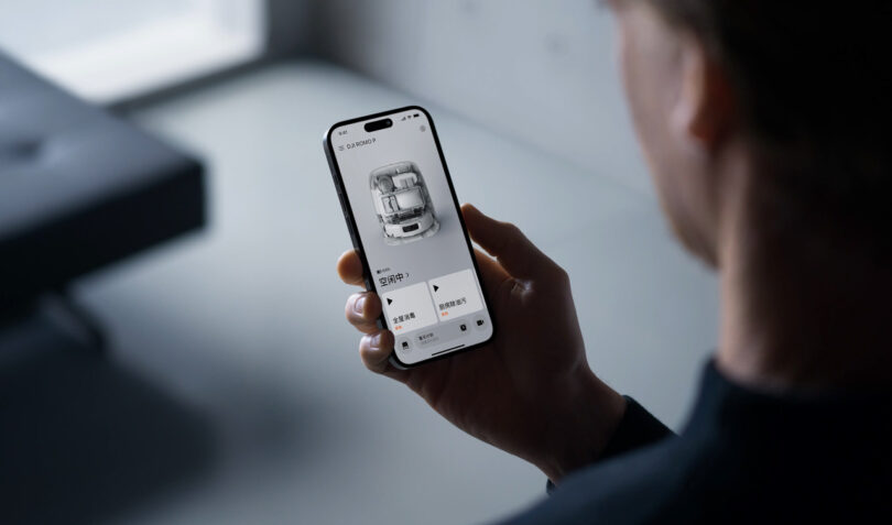 A person holds a smartphone displaying a car control app interface, featuring an overhead view of the car and various control options, similar in style to DJI Romo’s intuitive remote-control technology.