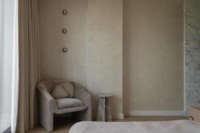 A minimalist bedroom corner with a beige armchair, small round side table, and three metallic wall accents on a light-colored textured wall.
