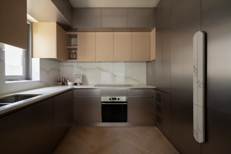 Minimalist modern kitchen with light wood cabinets, stainless steel surfaces, built-in oven, marble backsplash, and herringbone wood flooring.