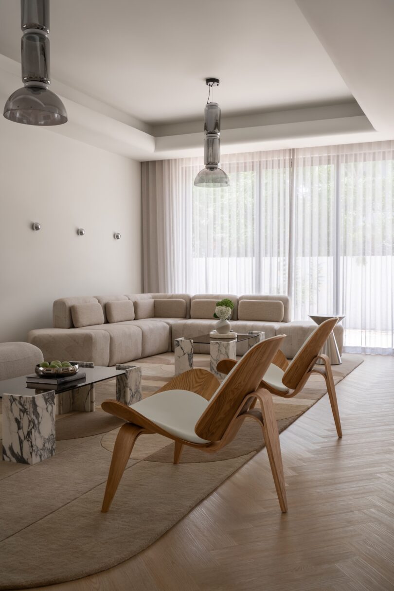 Modern living room with neutral tones, light wood chairs, a beige sectional sofa, marble tables, and floor-to-ceiling sheer curtains allowing natural light into the space.