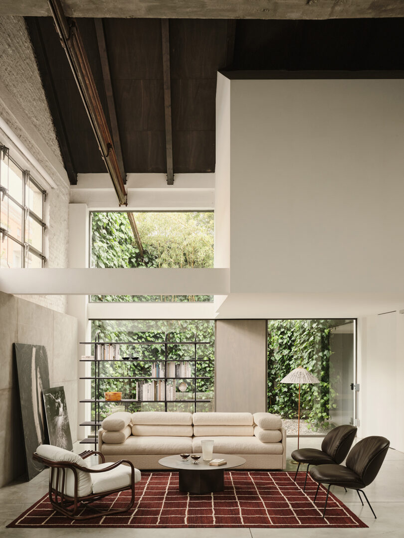 Modern living room with high ceilings, large windows, a beige sofa, two dark chairs, a round coffee table, a red patterned rug, and greenery visible outside.