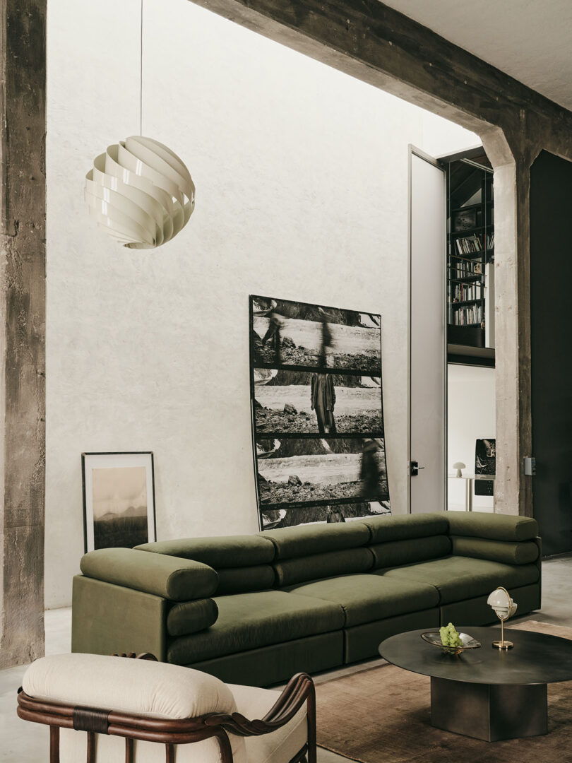 A modern living room with an olive-green sofa, a round coffee table, framed art leaning against the wall, and a pendant light hanging from the ceiling.