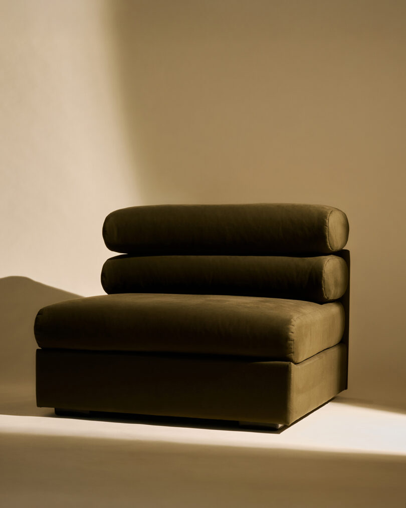 A modern, dark green armless lounge chair with three cylindrical cushions is placed on a light-colored floor against a beige background.