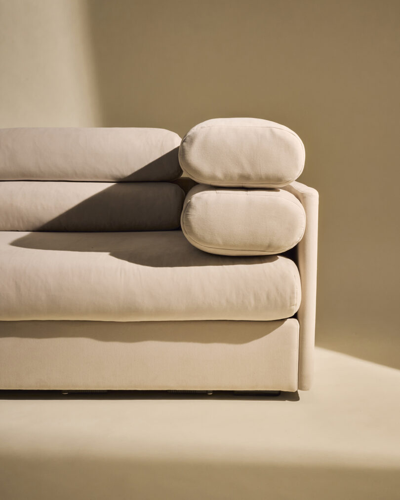 A close-up of a modern beige sofa with cylindrical cushions, shown against a neutral background with a diagonal shadow across the seat.