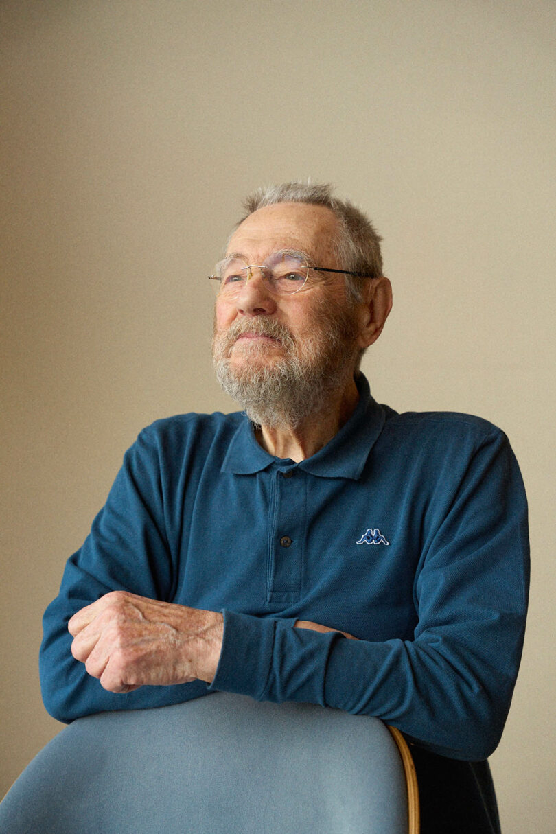 Elderly man with glasses and a gray beard sits behind a modern gray chair, wearing a dark blue long-sleeve polo, looking slightly upwards with a neutral expression.