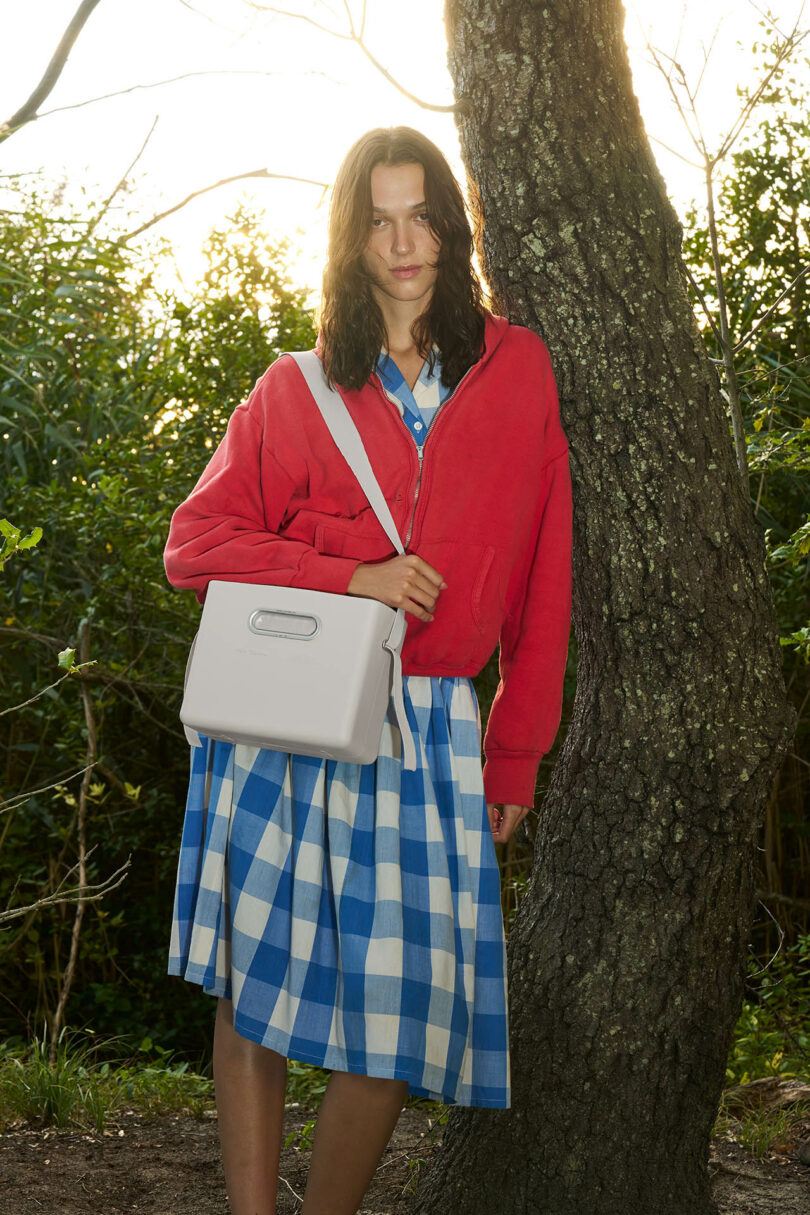 A person stands outdoors by a tree, wearing a red jacket, blue and white checkered dress, and carrying a light-colored shoulder bag.