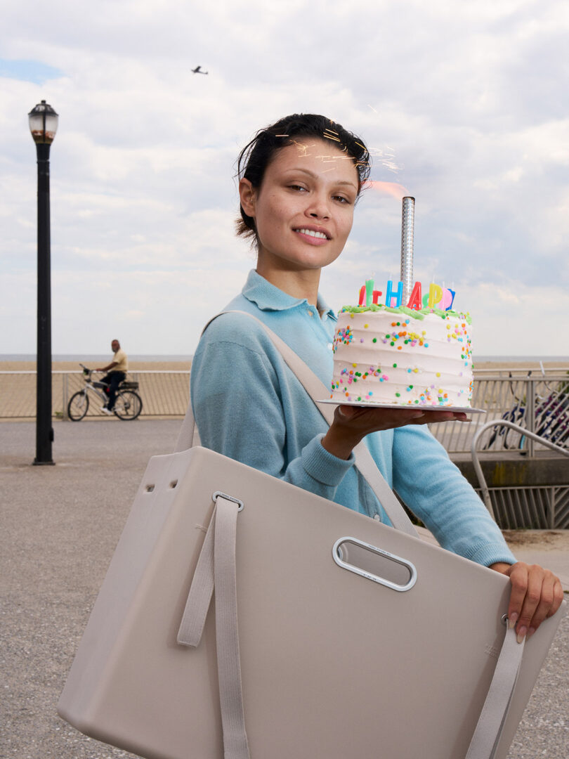 A person stands outdoors holding a birthday cake with candles and colorful sprinkles in one hand and carrying a large rectangular bag over their shoulder.