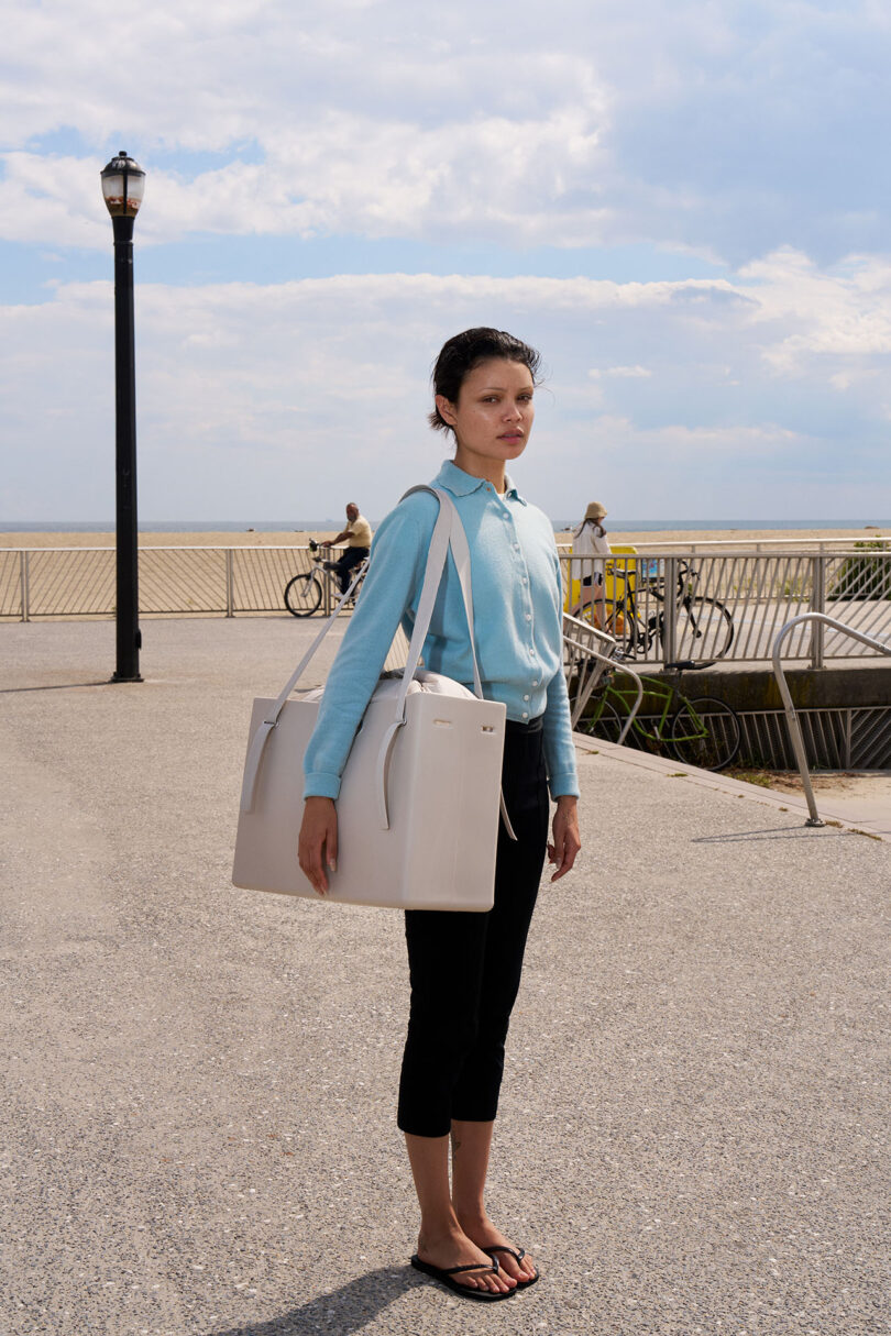 A person wearing a light blue shirt, black pants, and flip-flops stands outdoors holding a large white bag, with a beach and boardwalk in the background.