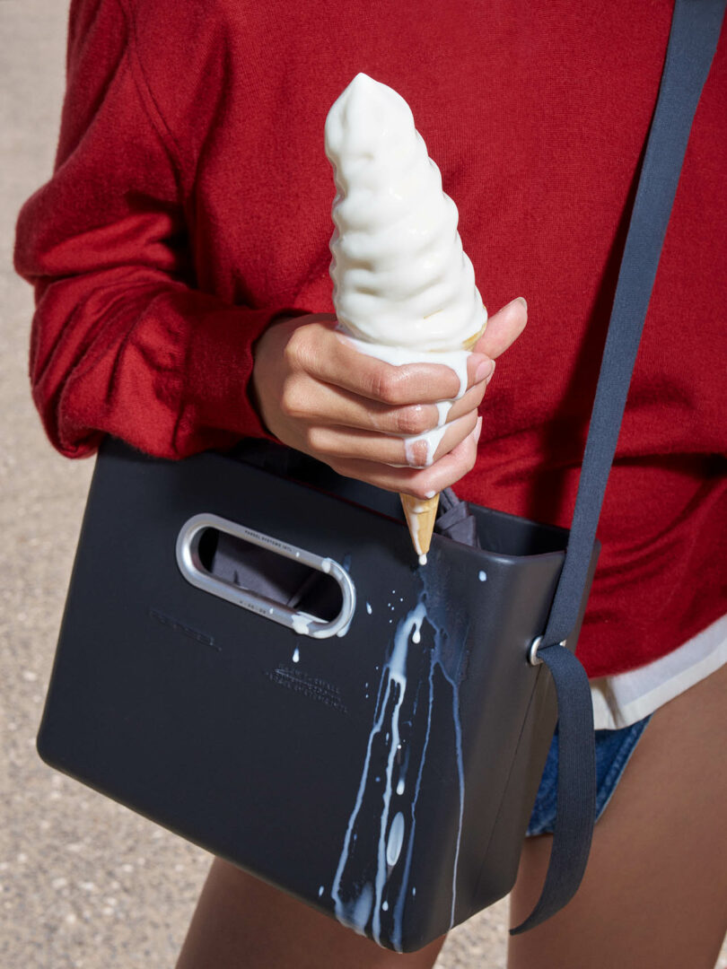 A person in a red sweater holding a melting soft-serve ice cream cone, with ice cream dripping onto a black bag.