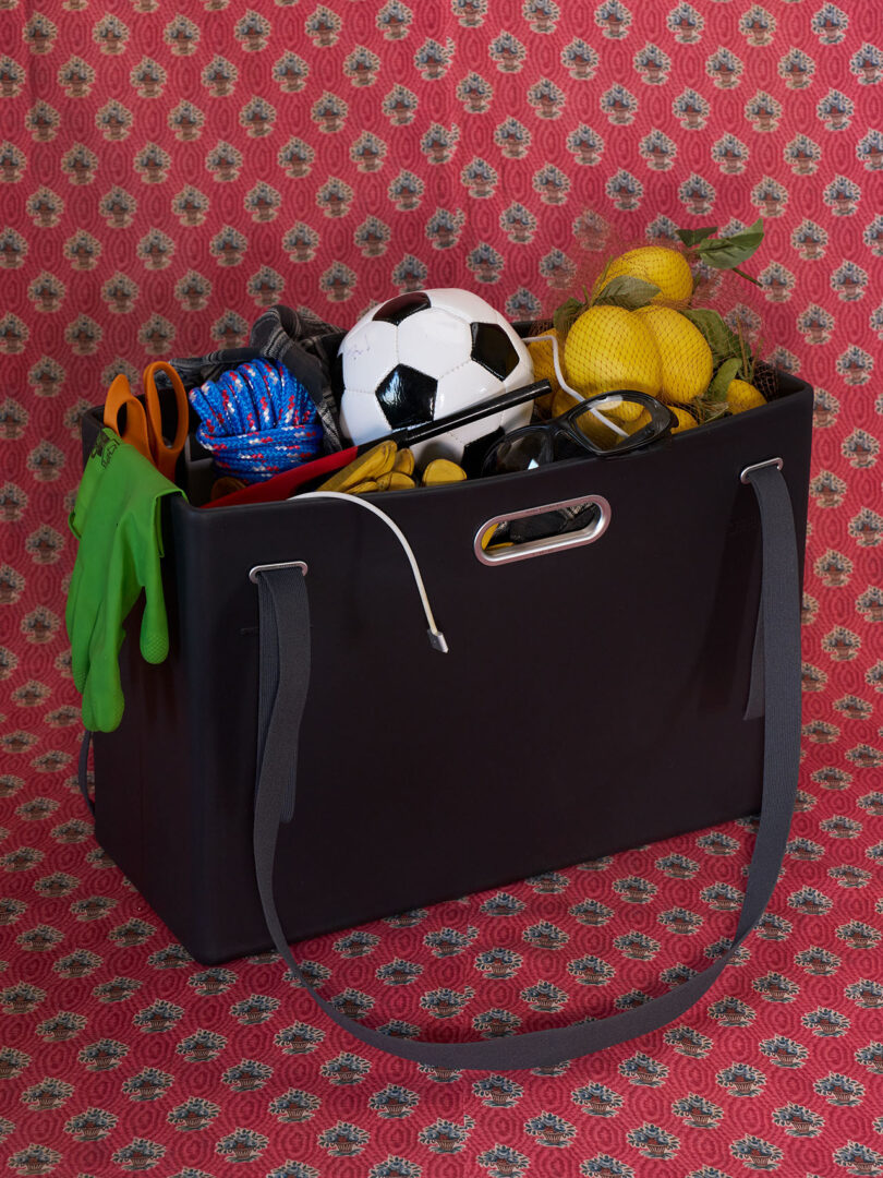 A black PARSEL EVA Tote System filled with a soccer ball, lemons, gardening gloves, scissors, rope, and other miscellaneous items sits in front of a red patterned background.