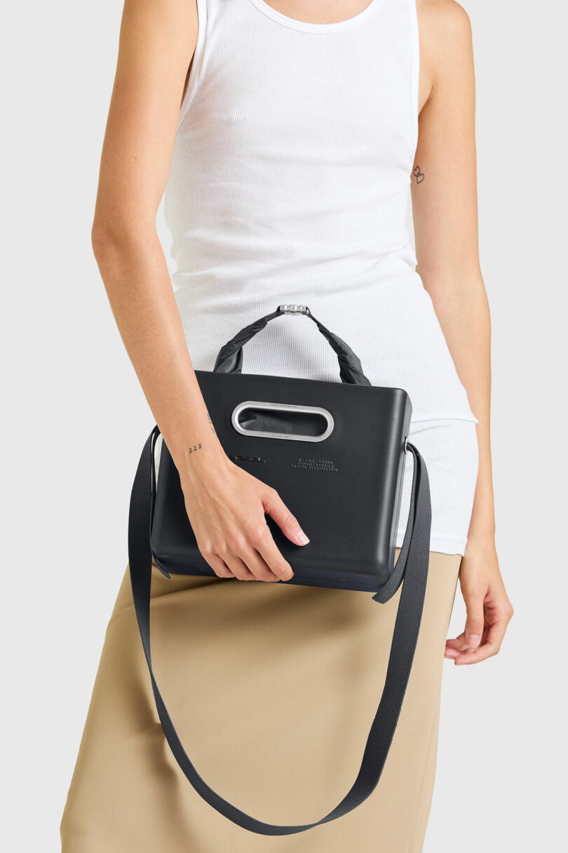 A person in a white sleeveless top and beige skirt holds a PARSEL EVA Tote System—a sleek black handbag with a handle and shoulder strap—against a plain background.