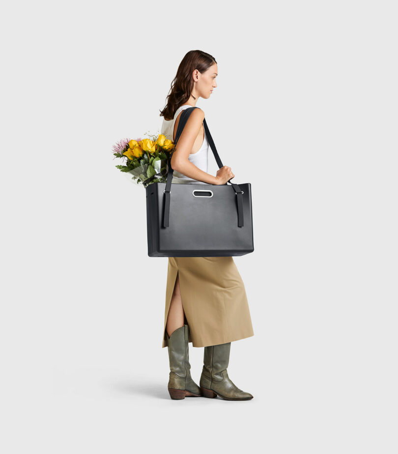 A woman in a white top, beige skirt, and boots carries the PARSEL EVA Tote System with yellow flowers inside, standing against a plain light background.
