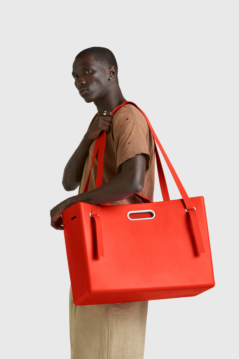 A person wearing a brown shirt and beige pants carries a large, rectangular, bright red PARSEL EVA Tote System bag with two long handles over their shoulder.