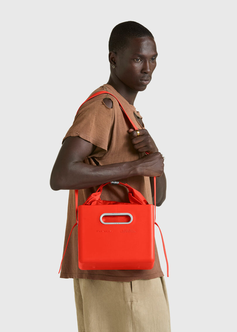 A person wearing a brown shirt and beige pants holds a bright red PARSEL EVA Tote System handbag with a shoulder strap against a plain light background.