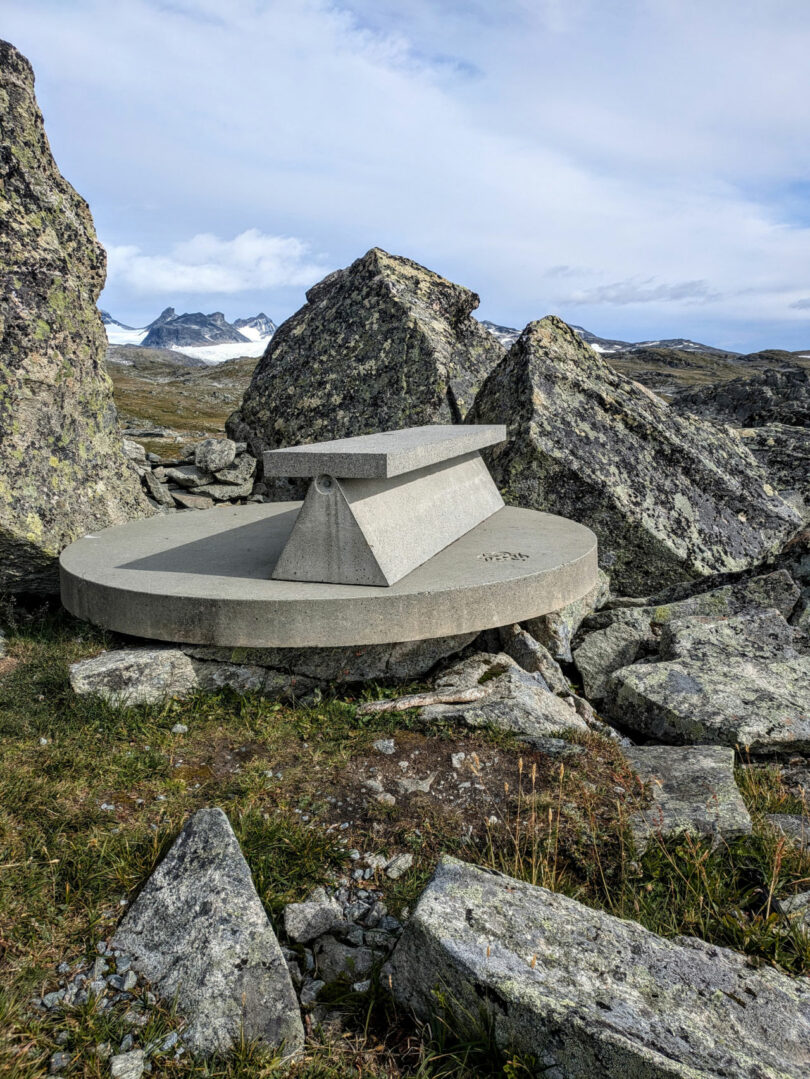 Concrete viewpoint marker with engraved information sits among large rocks on a mountainous landscape under a partly cloudy sky. Snow-capped peaks rise in the background, offering a breathtaking view from this Dana Harel-inspired setting.