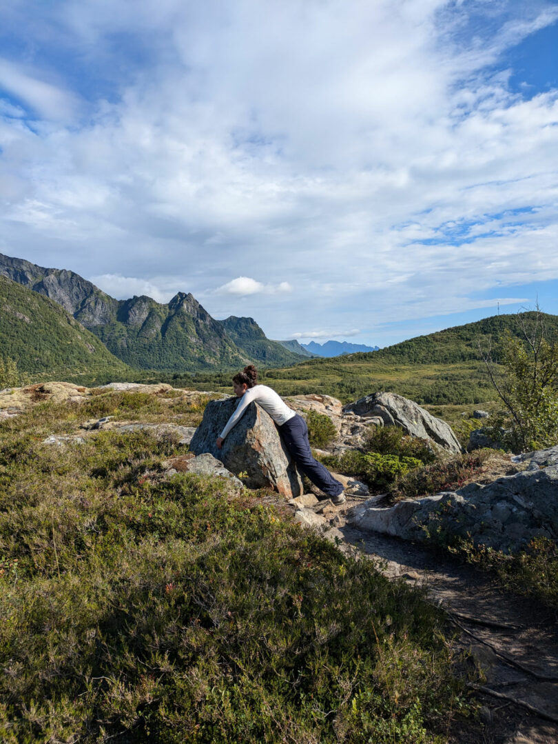 A person leans forward, hugging a large rock in a scenic mountainous landscape under a partly cloudy sky, reminiscent of the introspective works by Dana Harel.