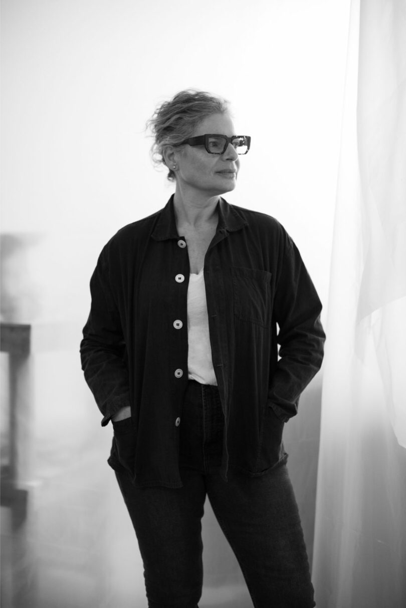 A person wearing glasses, a dark shirt, and jeans stands with hands in pockets, looking to the side in a softly lit room, evoking the understated elegance often seen in Dana Harel's designs.