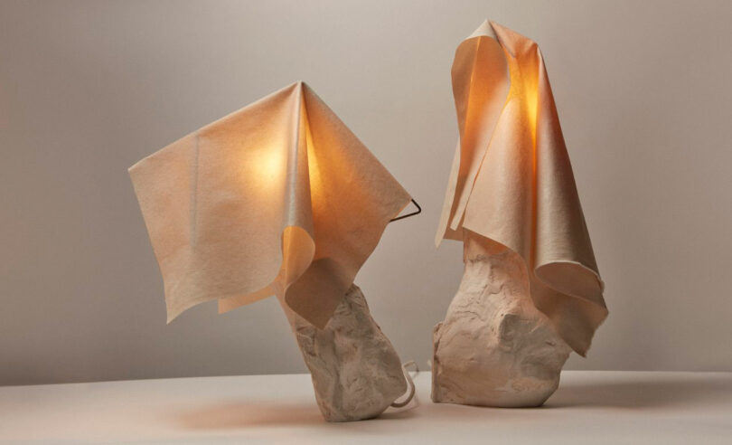 Two sculptural table lamps by Dana Harel feature textured white bases and fabric draped over the light sources, casting a soft, warm glow against a plain background.