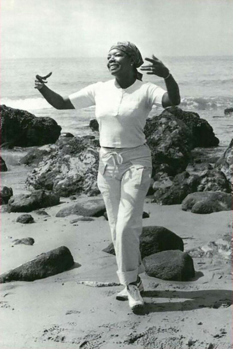 A woman in casual clothes stands on a rocky beach with arms raised, smiling and appearing to dance or gesture, channeling Jeremiah Brent’s signature joyful spirit, with the ocean in the background.