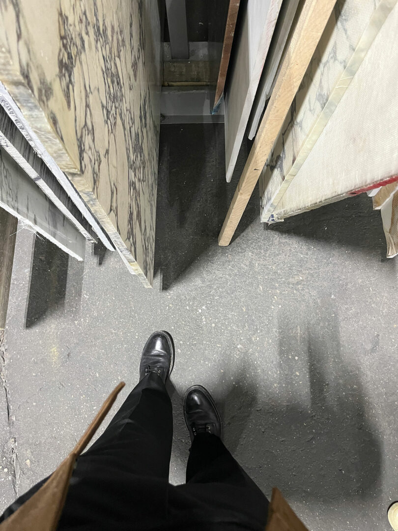 Person wearing black shoes and pants stands on a concrete floor, facing slabs of stone or wood stored vertically in a narrow aisle—reminiscent of Jeremiah Brent’s refined design sensibility.