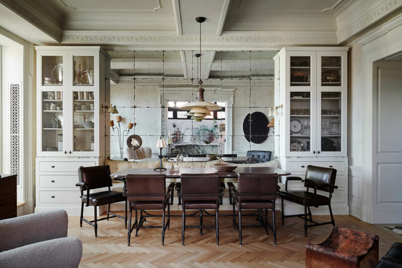 A Jeremiah Brent-inspired dining room features a wooden table, six chairs, built-in glass cabinets, mirrored wall, pendant light, and decorative items on display.