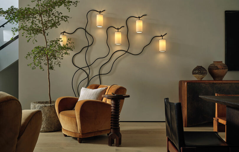 A modern living room inspired by Jeremiah Brent, featuring tan armchairs, a potted tree, a small side table, and five wall-mounted lights with artistic black wiring.