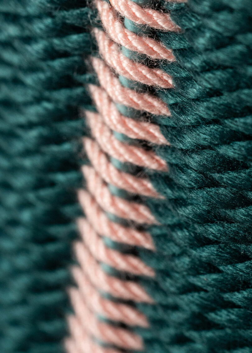 Close-up of teal and pink yarn, interwoven in a textured, diagonal stitch pattern inspired by Laura Casañas Maya.