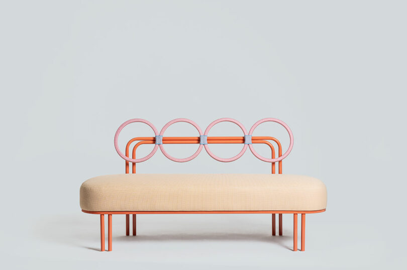 A modern bench by Laura Casañas Maya with a beige cushion, orange frame, and a backrest featuring four connected pink circles against a plain light gray background.