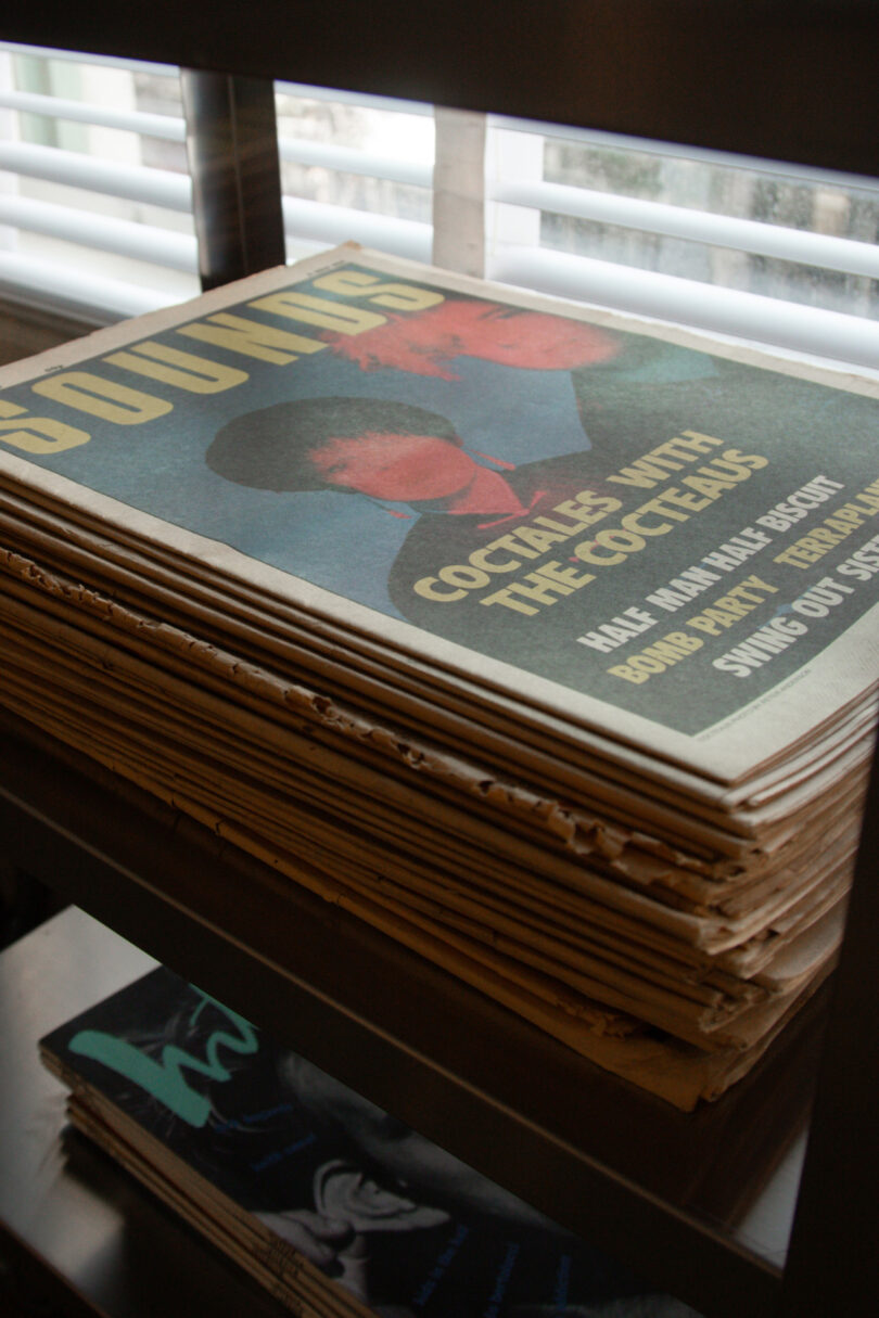 A stack of vintage magazines sits on a metal shelf near a window, with the top issue featuring a cover story titled "Cocktails with The Cocteaus."