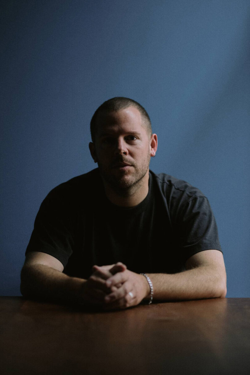 A man with short hair and a beard sits at a table with his hands clasped, in front of a plain blue background.