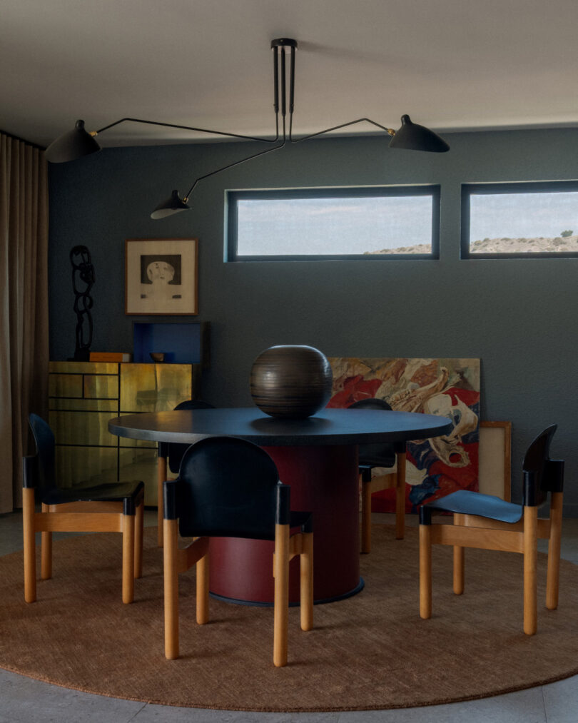 A modern dining area with a round table, four chairs, a dark centerpiece, wall art, and a sculptural ceiling light above a brown rug. Two rectangular windows provide natural light.