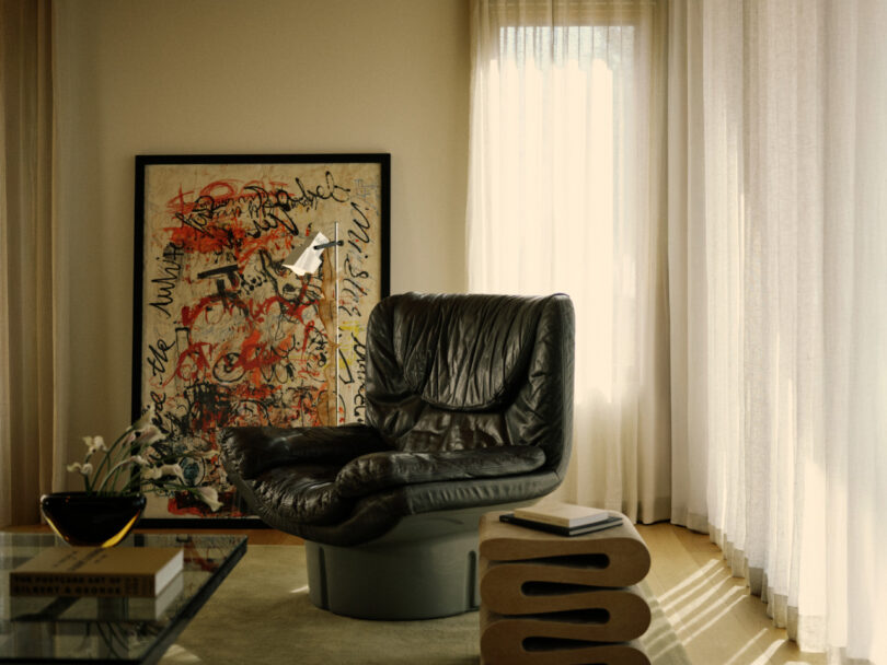 A modern living room with a black leather lounge chair, abstract framed artwork, sculptural side table, glass coffee table, and sheer curtains letting in natural light.