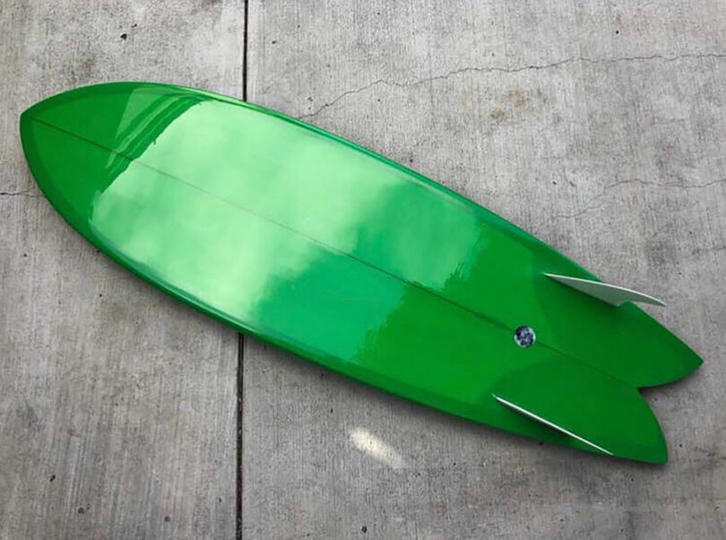 A green surfboard with twin fins and a swallowtail design lies on a concrete surface.