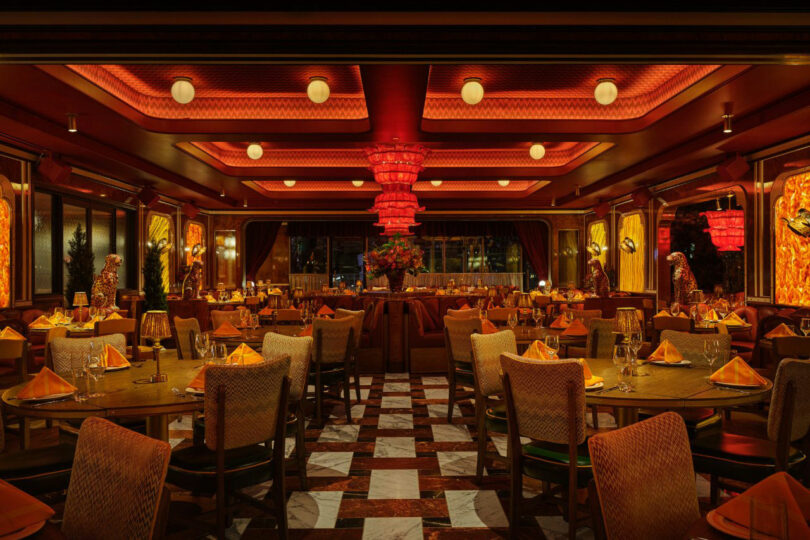 Restaurant interior with red lighting, chandeliers, neatly set tables with orange napkins, upholstered chairs, and a central floral arrangement. Marble-patterned floor tiles are visible.
