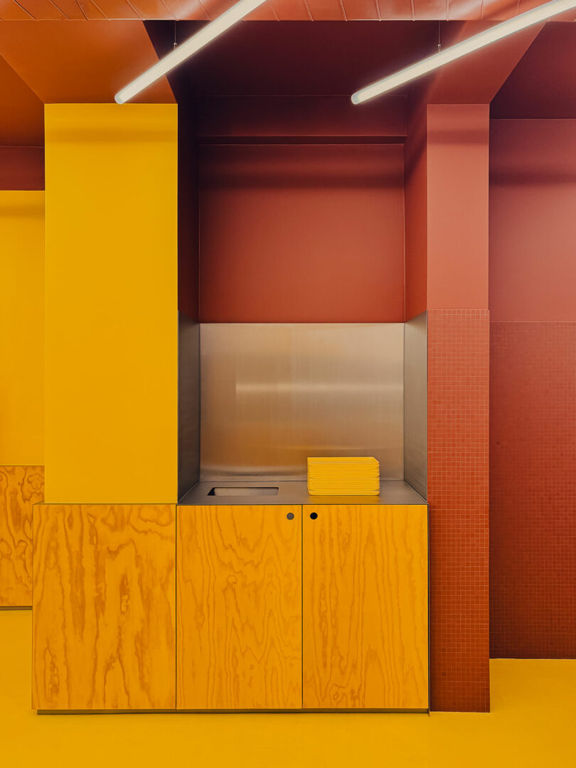 A modern kitchen area features yellow wood cabinets, a stainless steel backsplash, a stack of yellow plates, and red walls with overhead linear lights.