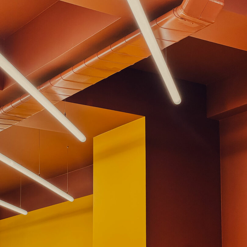 Ceiling with suspended linear LED lights, exposed orange ductwork, and intersecting yellow and dark red walls in a modern interior space.