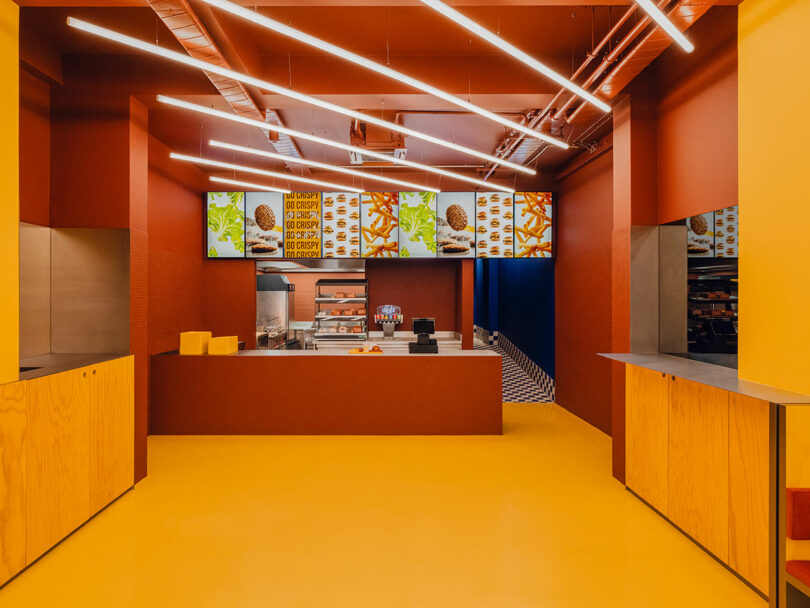 A brightly lit fast food counter with digital menu boards displaying burgers, fries, and drinks, featuring yellow floors and wooden paneling.