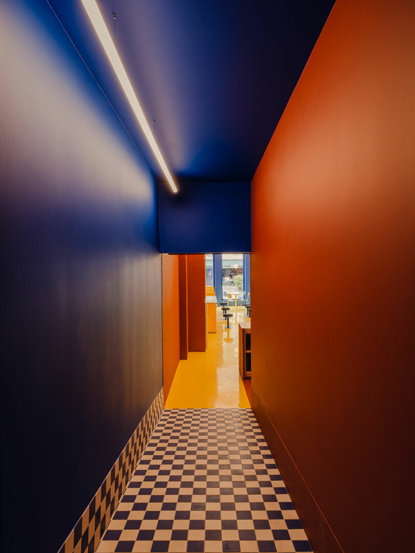 A hallway with blue and orange walls, a checkered black-and-white tile floor, and a view into a bright room with tables and chairs in the distance.