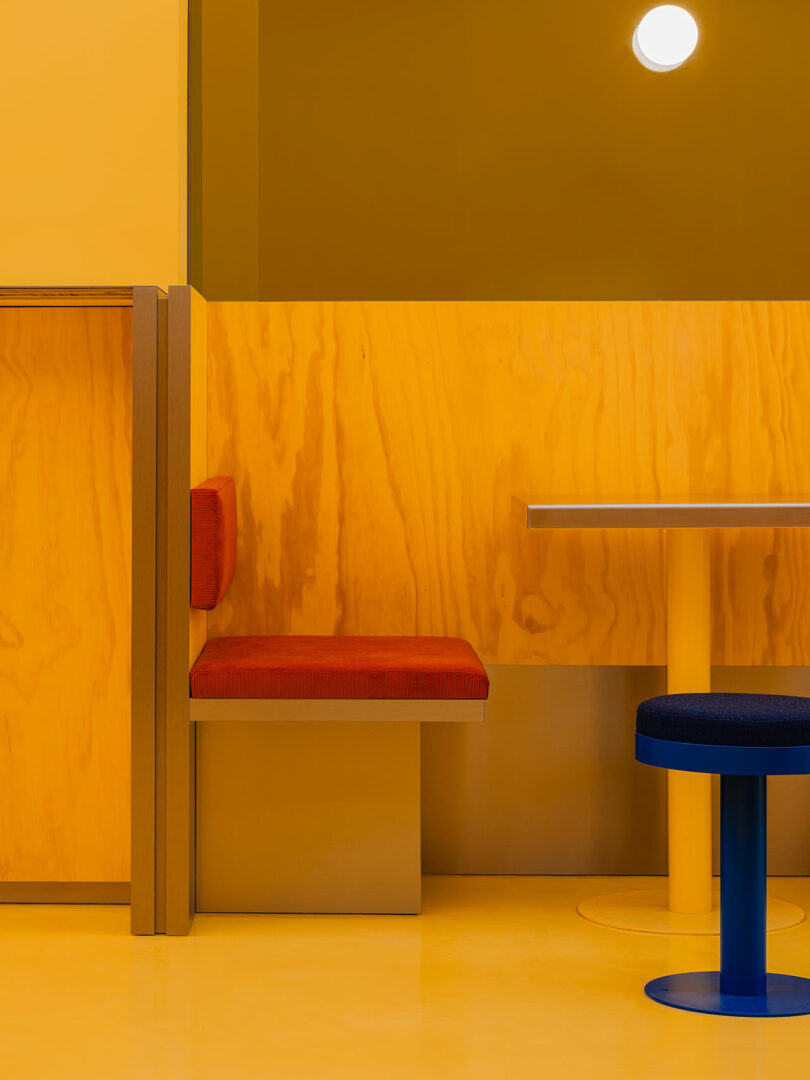 A minimalist cafe interior with yellow walls, a wooden booth with an orange cushion, a metal table, and a blue stool under a round wall light.