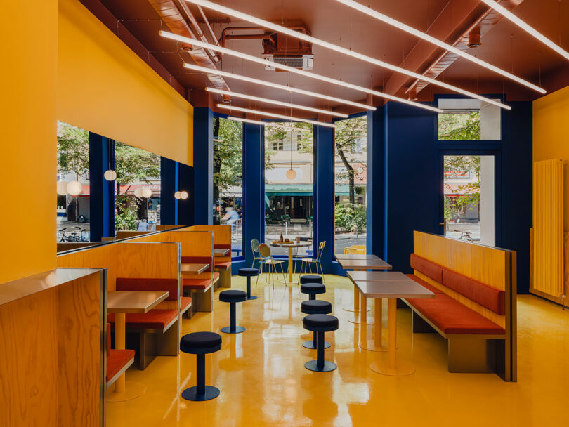 Modern cafe interior with yellow floors and walls, red ceiling, blue accents, wooden booths with red cushions, round tables, and large windows facing a street with trees.