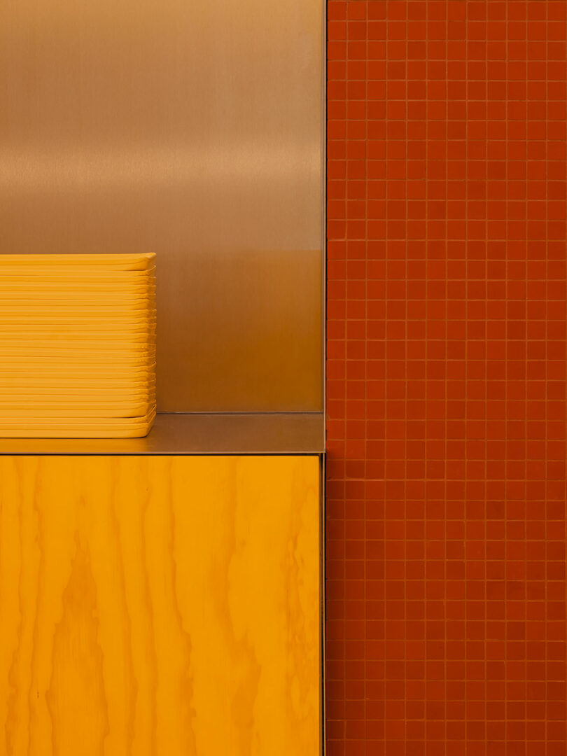 A stack of yellow trays sits on a metal counter with an orange wood cabinet below and a red tiled wall to the right.