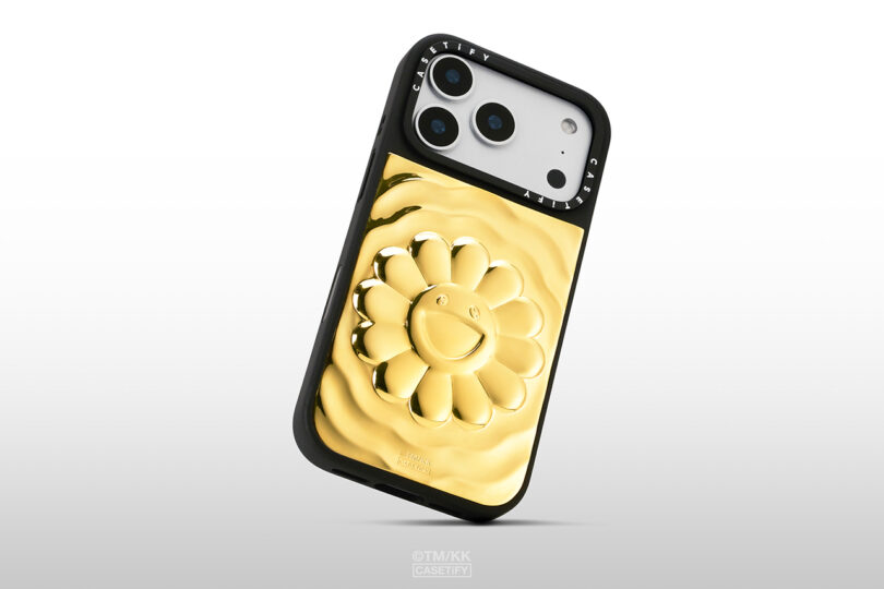 A smartphone with a black case featuring a raised, shiny gold flower design with a smiling face on the back.