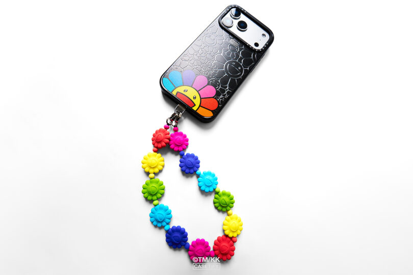A smartphone in a black case with a colorful smiling flower design and a detachable chain of multicolored flower beads attached.