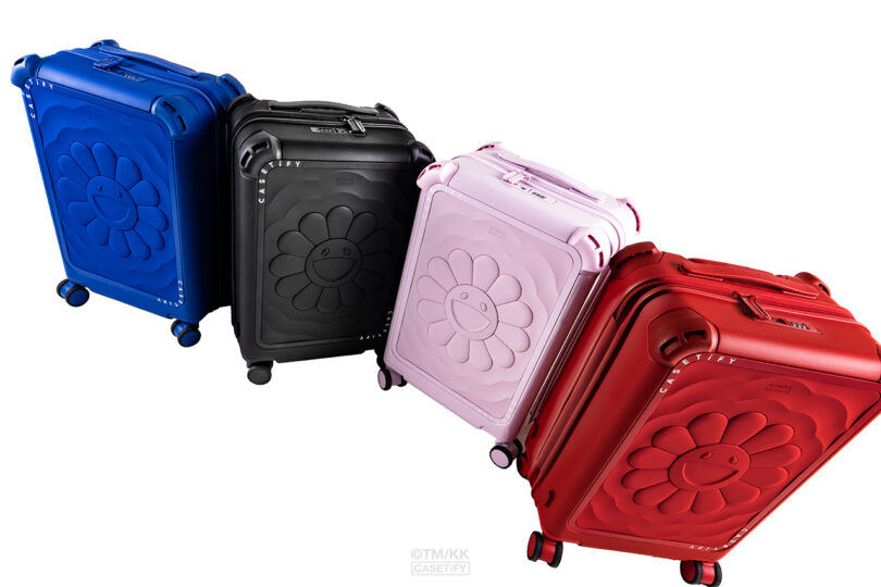 Four hard-shell suitcases in blue, black, pink, and red, each featuring a raised flower design with a smiley face, are displayed against a white background.