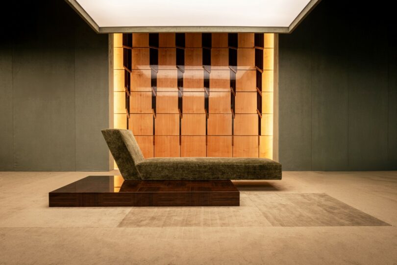 A modern, minimalist lounge chair with an angular design sits on a platform in front of a backlit wooden shelving wall in a contemporary room